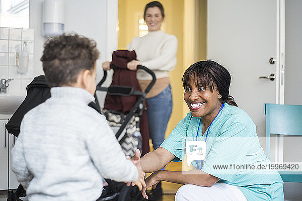 Smiling female nurse shaking hands with boy while mother standing in background at medical clinic
