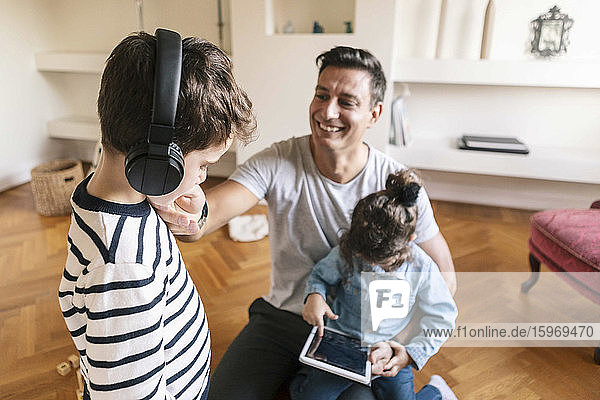 Lächelnder Vater sieht liebevollen Sohn an  während die Tochter zu Hause ein digitales Tablet benutzt