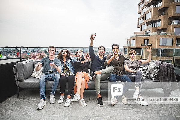 Porträt von lächelnden Freunden und Freundinnen mit Getränken  die auf dem Sofa auf der Gebäudeterrasse für geselliges Beisammensein sitzen