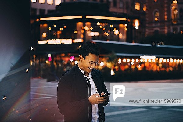 Smiling young man using mobile phone while standing in illuminated city at night