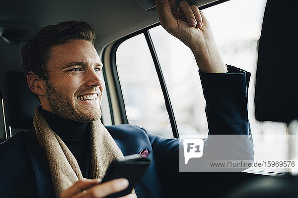 Smiling businessman with phone looking away while sitting in taxi