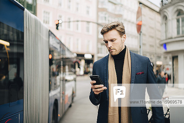 Male entrepreneur using internet through mobile phone while standing by bus in city