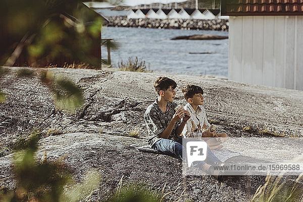Male siblings looking away while sitting on blanket over archipelago during weekend