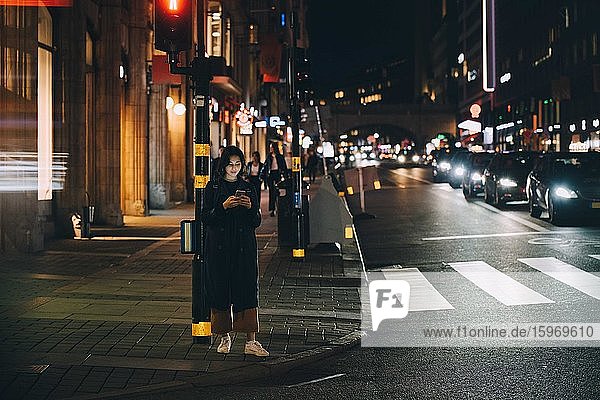 Frau in voller Länge  die ein Smartphone benutzt  während sie nachts auf einem Fußweg in der Stadt steht