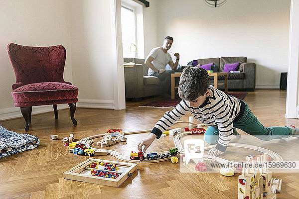 Sohn spielt mit Miniatur-Eisenbahn  während der Vater zu Hause im Hintergrund auf dem Sofa sitzt