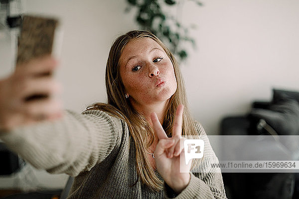 Teenager-Mädchen zeigt Friedenszeichen  während sie sich zu Hause über das Mobiltelefon selbstständig macht