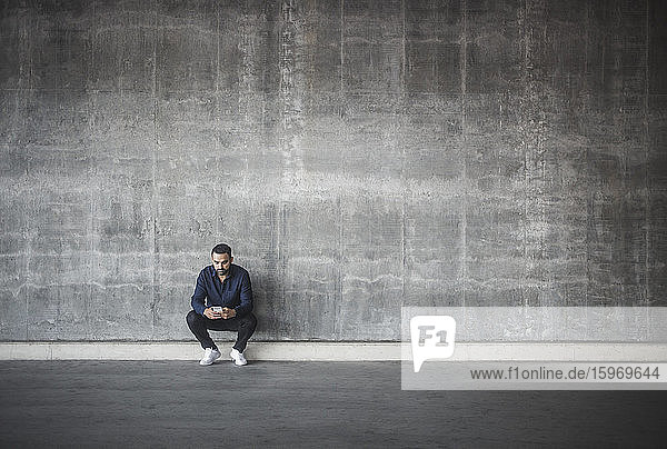 Contemplating man using cell phone while crouching against gray wall