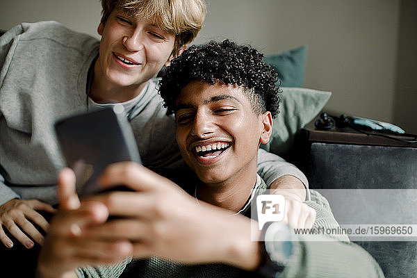 Lächelnder Teenager-Junge verschickt Textnachrichten über ein Smartphone  während er mit einem Freund zu Hause sitzt