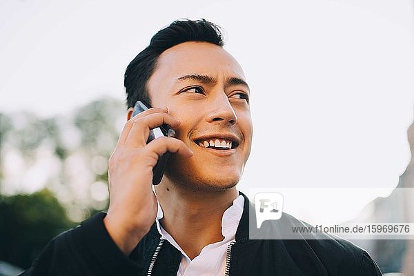 Smiling man talking through smart phone while looking away against sky
