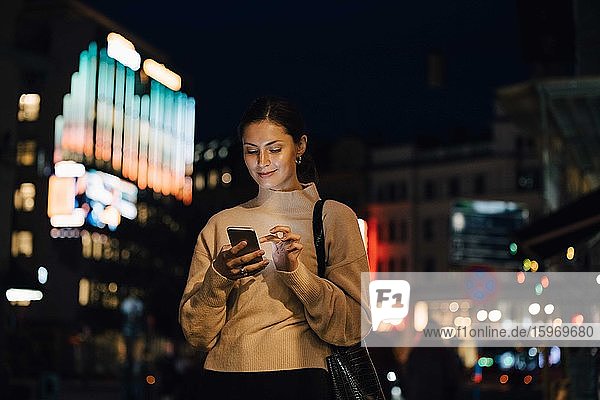 Lächelnde Frau schreibt nachts in der Stadt per Smartphone SMS