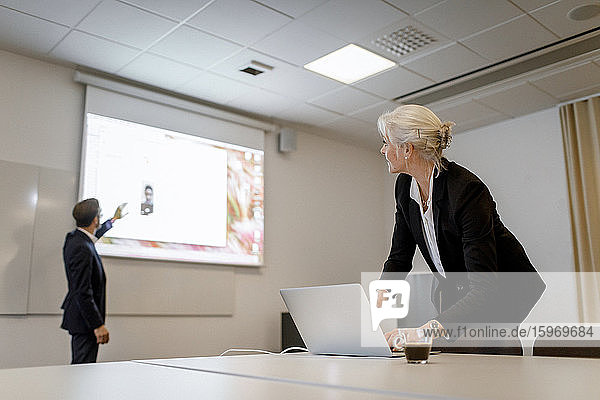 Eine weibliche Fachkraft benutzt einen Laptop  während ein männlicher Kollege während einer Videokonferenz im Sitzungssaal bei o