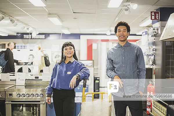 Portrait of smiling salesman and saleswoman standing by luggage cart in electronics store