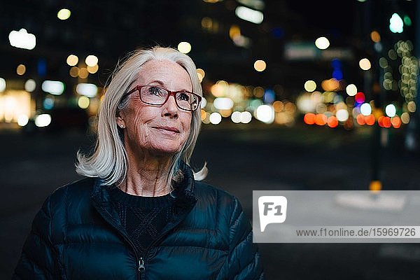 Faltig lächelnde Frau schaut auf  während sie nachts in einer beleuchteten Stadt steht