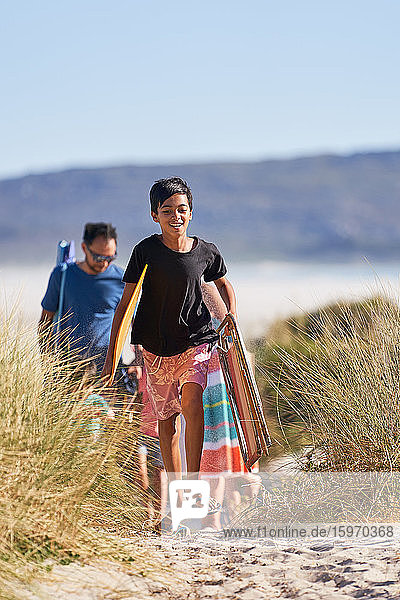 Glücklicher Junge trägt Klappstuhl und Handtuch am sonnigen Strand