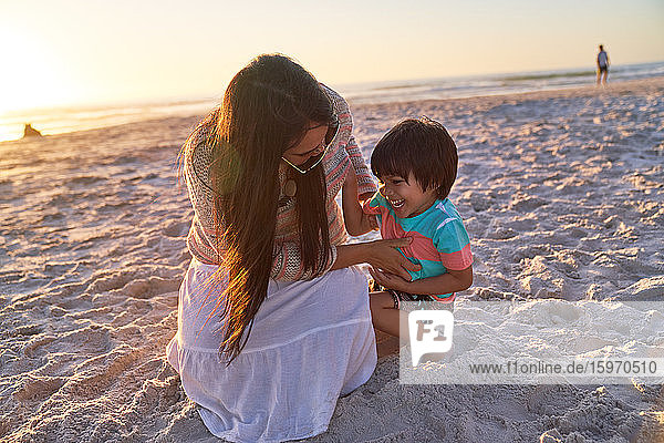 Verspielte Mutter kitzelt Sohn am Strand bei Sonnenuntergang