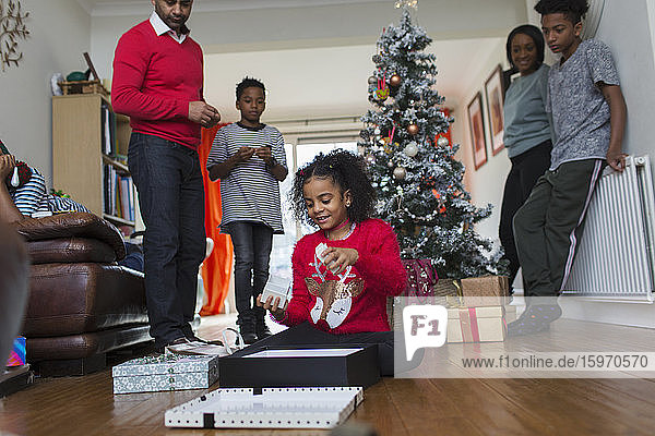 Familie beobachtet Mädchen beim Öffnen von Weihnachtsgeschenken auf dem Wohnzimmerboden