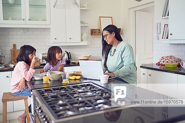 Mother working at laptop while daughters eat breakfast in kitchen