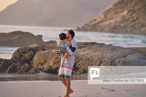 Affectionate father hugging and kissing son on ocean beach