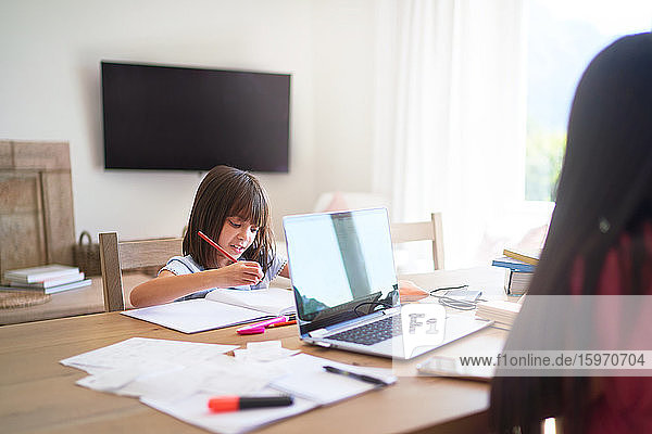 Mädchen macht Hausaufgaben  während die Mutter am Laptop am Esstisch arbeitet