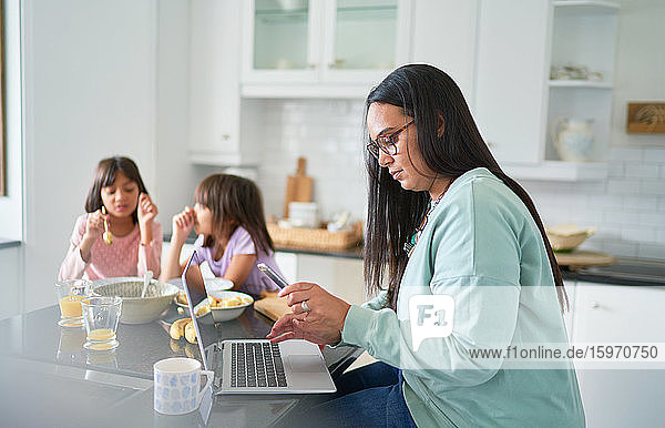 Mutter arbeitet am Laptop in der Küche  während die Töchter frühstücken