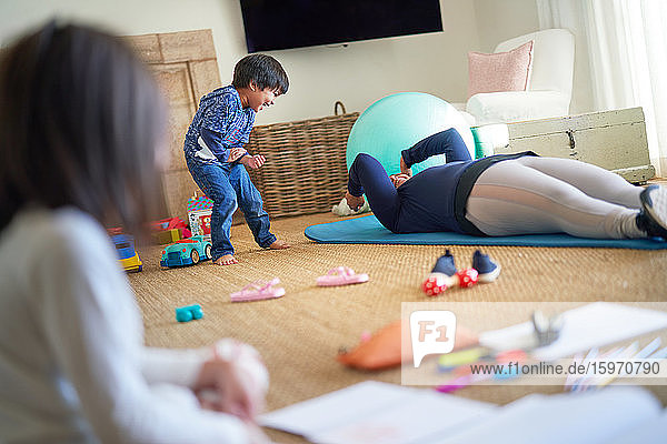 Verspielter Junge schaut der Mutter beim Training auf dem Wohnzimmerboden zu