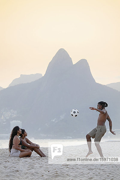Ein junger schwarzer Brasilianer spielt mit zwei Freundinnen Fussball am Strand von Ipanema mit dem Dois Irmaos-Gebirge in der Ferne  Rio de Janeiro  Brasilien  Südamerika