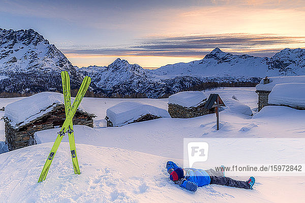 Junger Skifahrer entspannt sich im Schnee mit Blick auf das kleine Dorf bei Sonnenaufgang  Valmalenco  Valtellina  Lombardei  Italien  Europa