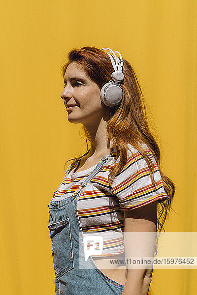 Smiling young woman listening music though headphones against yellow wall during sunny day
