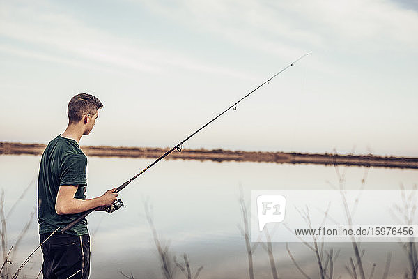 Side view of teenage boy fishing with rod in lake while standing against cloudy sky