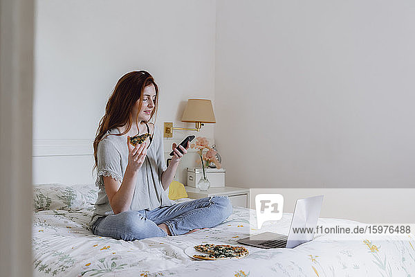Beautiful woman using mobile phone while sitting with pizza and laptop on bed at home