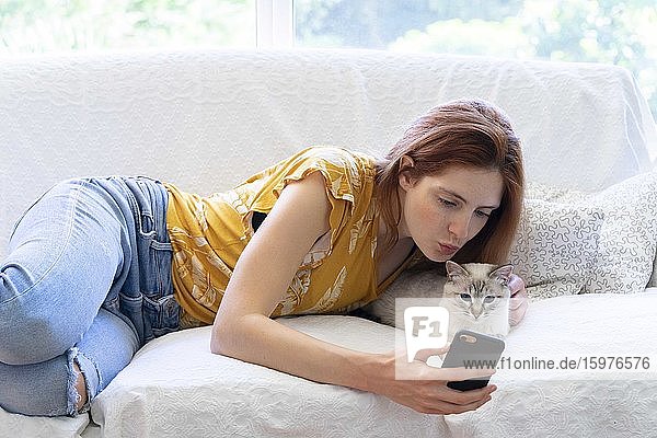 Junge Frau  die mit ihrer Katze zu Hause auf der Couch liegt und ihr Smartphone benutzt