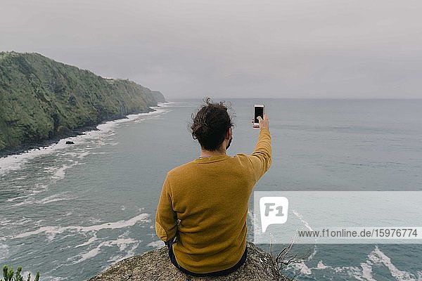 Rear view of man sitting on a rock at the coast taking a smartphone picture on Sao Miguel Island  Azores  Portugal