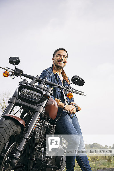 Smiling young biker leaning on motorcycle against clear sky