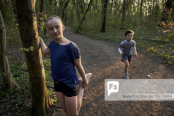 Mädchen schaut weg  während sie ihr Bein am Baumstamm gegen den im Wald laufenden Bruder streckt