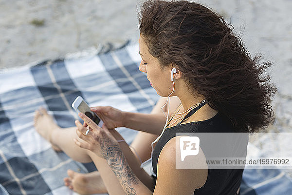 Junge Frau sitzt auf einer Decke am Strand und benutzt Kopfhörer und Smartphone