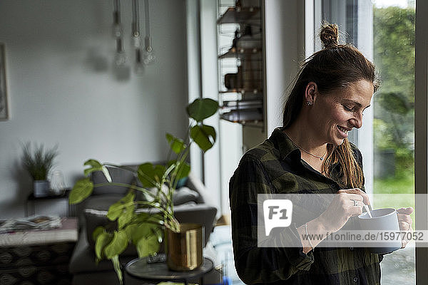Lächelnde erwachsene Frau  die einen Kaffeebecher hält  während sie zu Hause am Fenster steht