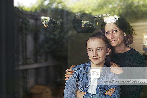Lächelnde Frau und Tochter schauen durch das Fenster  während sie zu Hause stehen