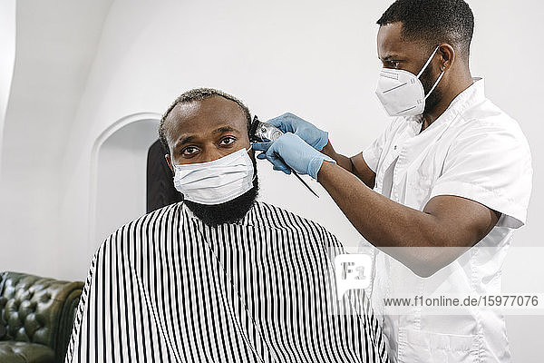 Friseur mit chirurgischer Maske und wiederverwendbaren Handschuhen beim Rasieren der Haare eines Kunden