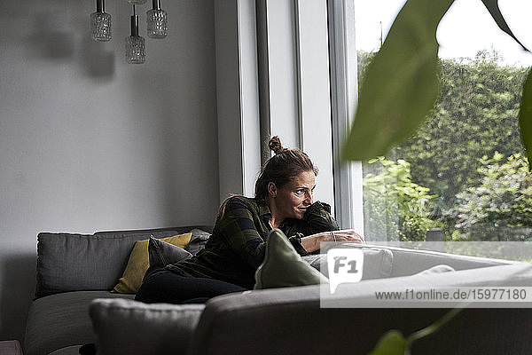 Nachdenkliche Frau schaut durch das Fenster  während sie sich zu Hause auf dem Sofa entspannt