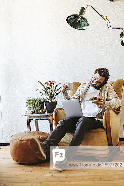 Lächelnder Mann am Telefon auf einem Sessel zu Hause mit Laptop  Croissant und einer Tasse Kaffee