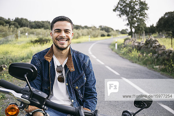 Lächelnder junger Motorradfahrer auf dem Motorrad sitzend auf dem Lande