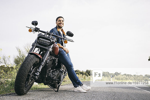 Lächelnder junger Motorradfahrer lehnt auf dem Motorrad auf der Straße gegen den Himmel