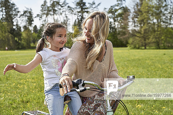 Lächelnde blonde Frau sieht ihre Tochter an  die mit ausgestreckten Armen Fahrrad fährt