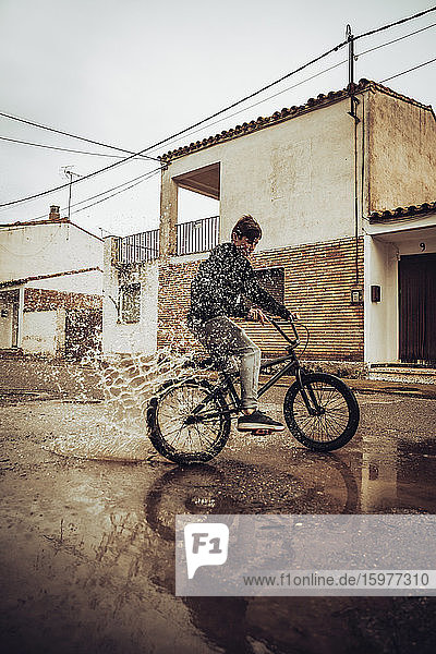 Jugendlicher spritzt Wasser in eine Pfütze  während er in der Regenzeit auf der Straße Fahrrad fährt
