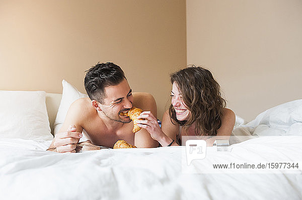 Glückliche junge Frau füttert einen Mann mit einem Croissant  während sie zu Hause im Bett liegt