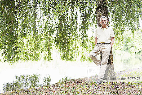 Porträt eines lächelnden älteren Mannes  der sich an einen Baumstamm in einem Park lehnt