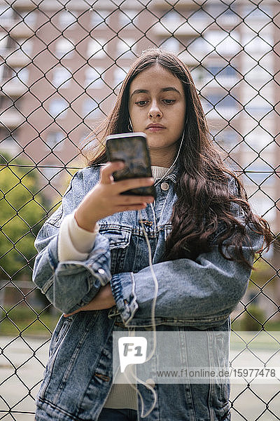Confident teenage girl using smart phone against chainlink fence at sports court