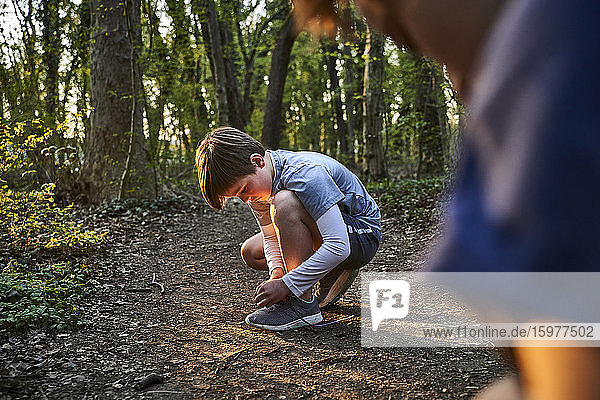 Seitenansicht eines Jungen in voller Länge  der sich beim Binden von Schnürsenkeln im Wald hinhockt