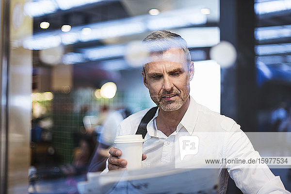 Businessman holding coffee reading newspaper while sitting in train seen through window