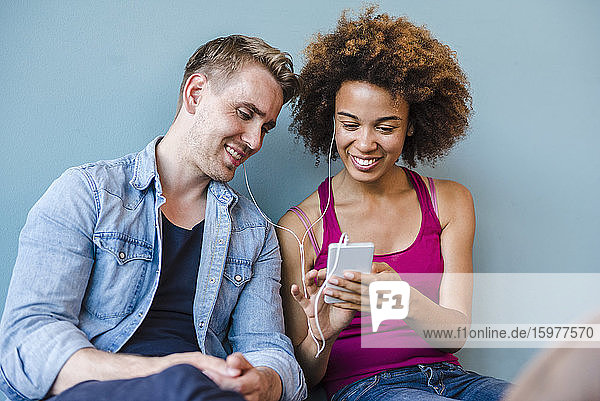 Junge Frau und Mann sitzen in einem modernen Büro auf dem Boden  teilen sich Kopfhörer und benutzen ein Smartphone